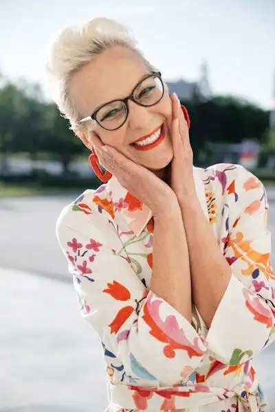 Smiling older woman with short blonde hair wearing glasses and a colorful floral dress, standing outdoors with her hands gently cupping her face.